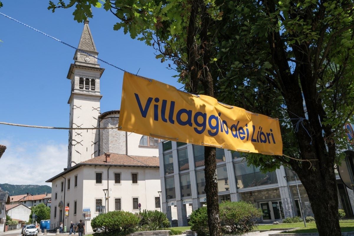 Striscione con la scritta «Villaggio dei Libri» appeso tra gli alberi, con sullo sfondo il campanile di una chiesa